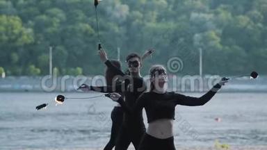 有技巧的男人和两个穿着黑色衣服和面具的女人站在河岸上用火焰<strong>表演节目</strong>。 技能